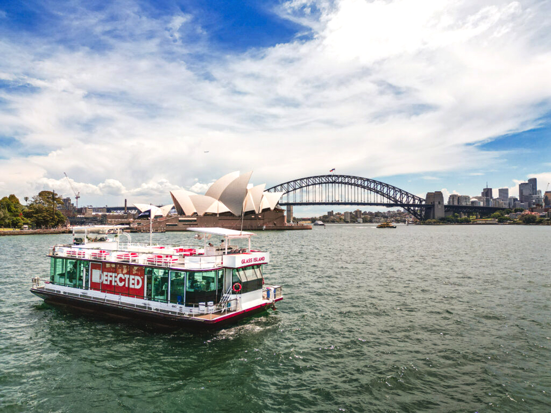 Glass Island passenger boat sails on Sydney Harbour beneath a blue sky, with the Opera House and Harbour Bridge in the background.